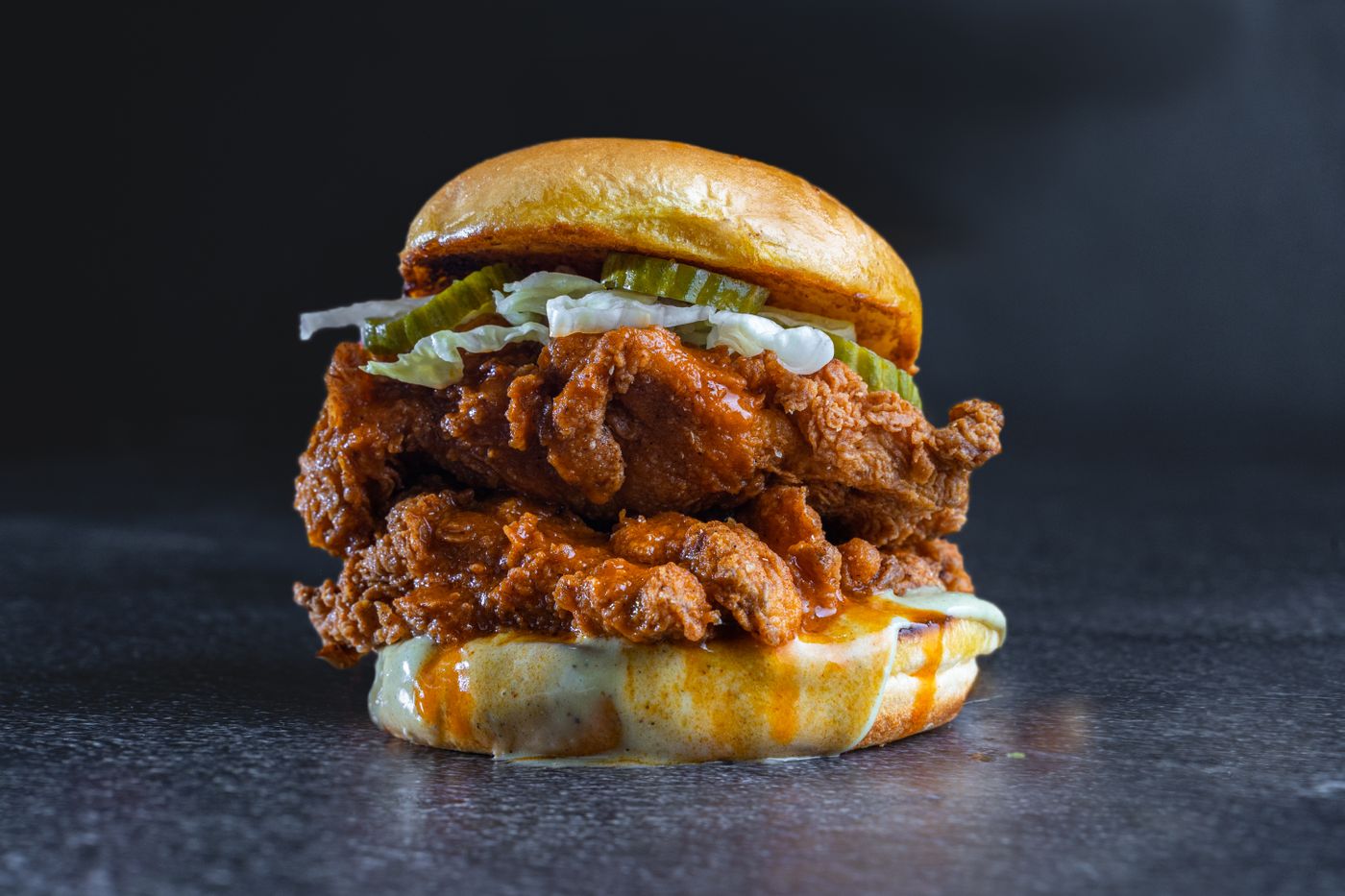 Crispy chicken burger with pickles. breaded fried chicken; round glazed bun; pickle slices; pale shredded lettuce; melted pale cheese; creamy sauce.