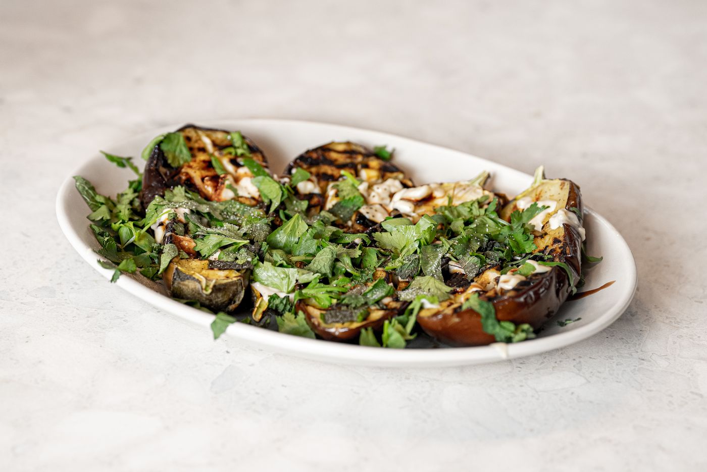 Herby roasted eggplant with tahini; roasted eggplant rounds, chopped fresh green herbs, white creamy drizzle, browned nuts or seeds