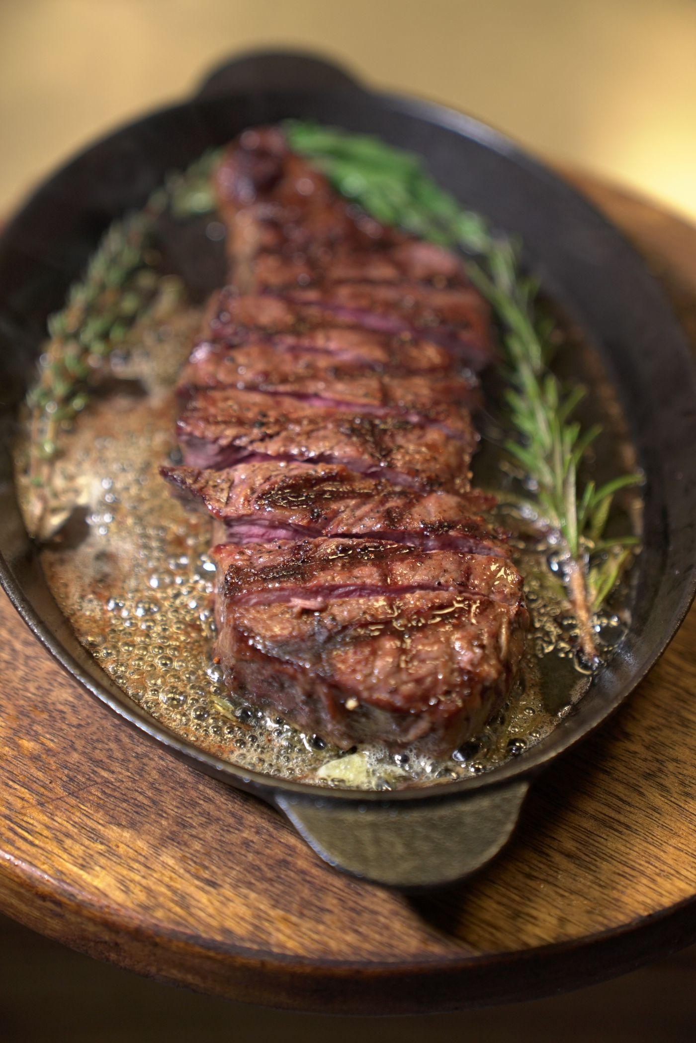 Sizzling rosemary steak. long seared steak; dark grill marks; rosemary sprigs; bubbling golden pan fat.