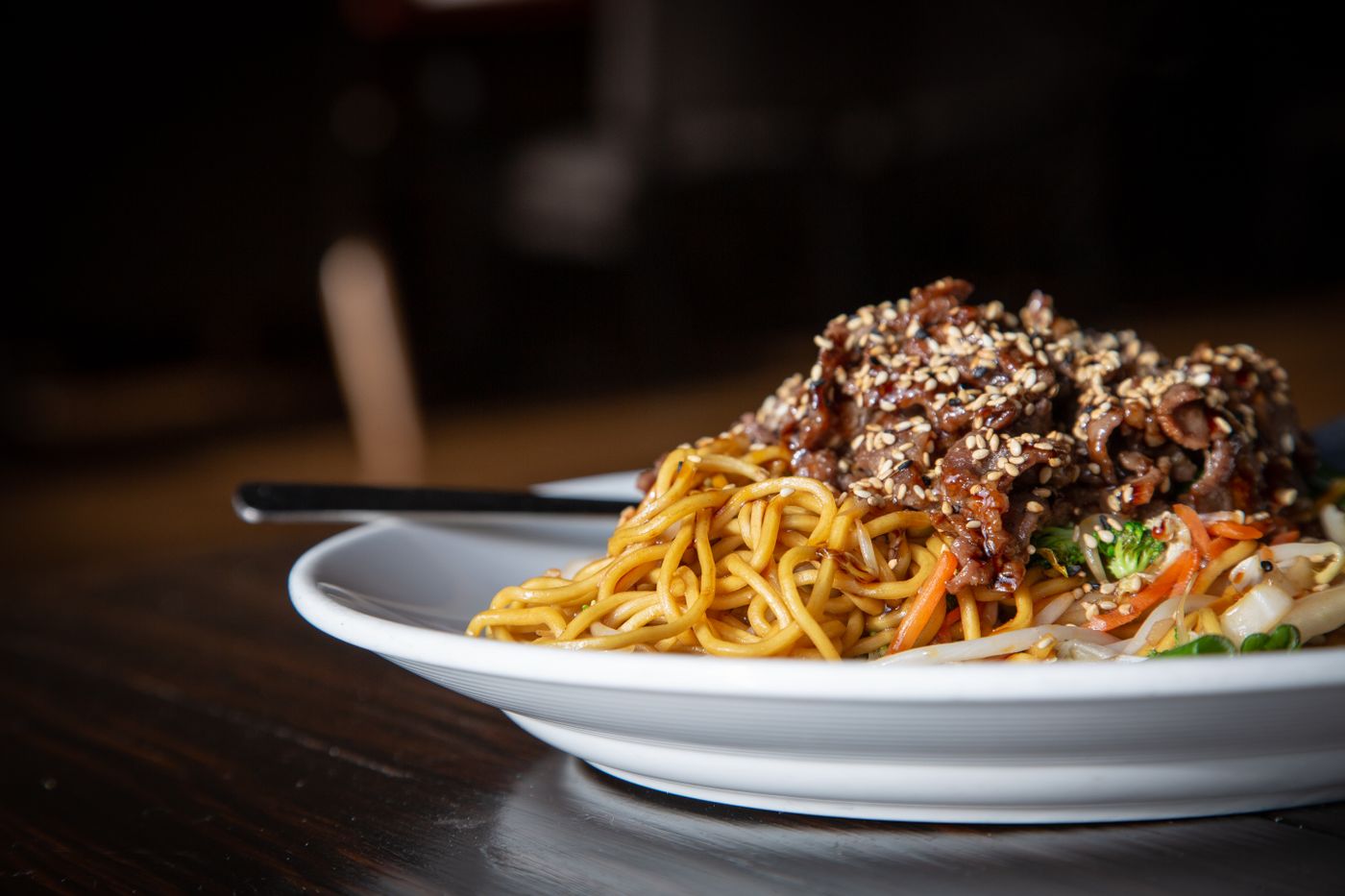 Sesame beef noodles with crunchy vegetables. thin yellow noodles; browned sliced beef; sesame seeds; carrot strips; shredded pale cabbage; green vegetables.