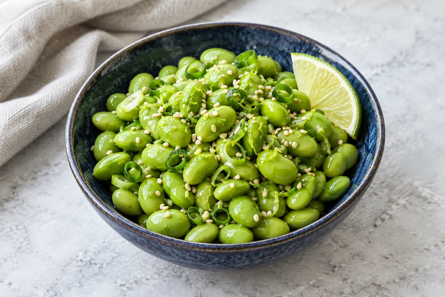 green shelled edamame with sesame seeds, scallion rings, lime zest and a lime wedge in a blue bowl.