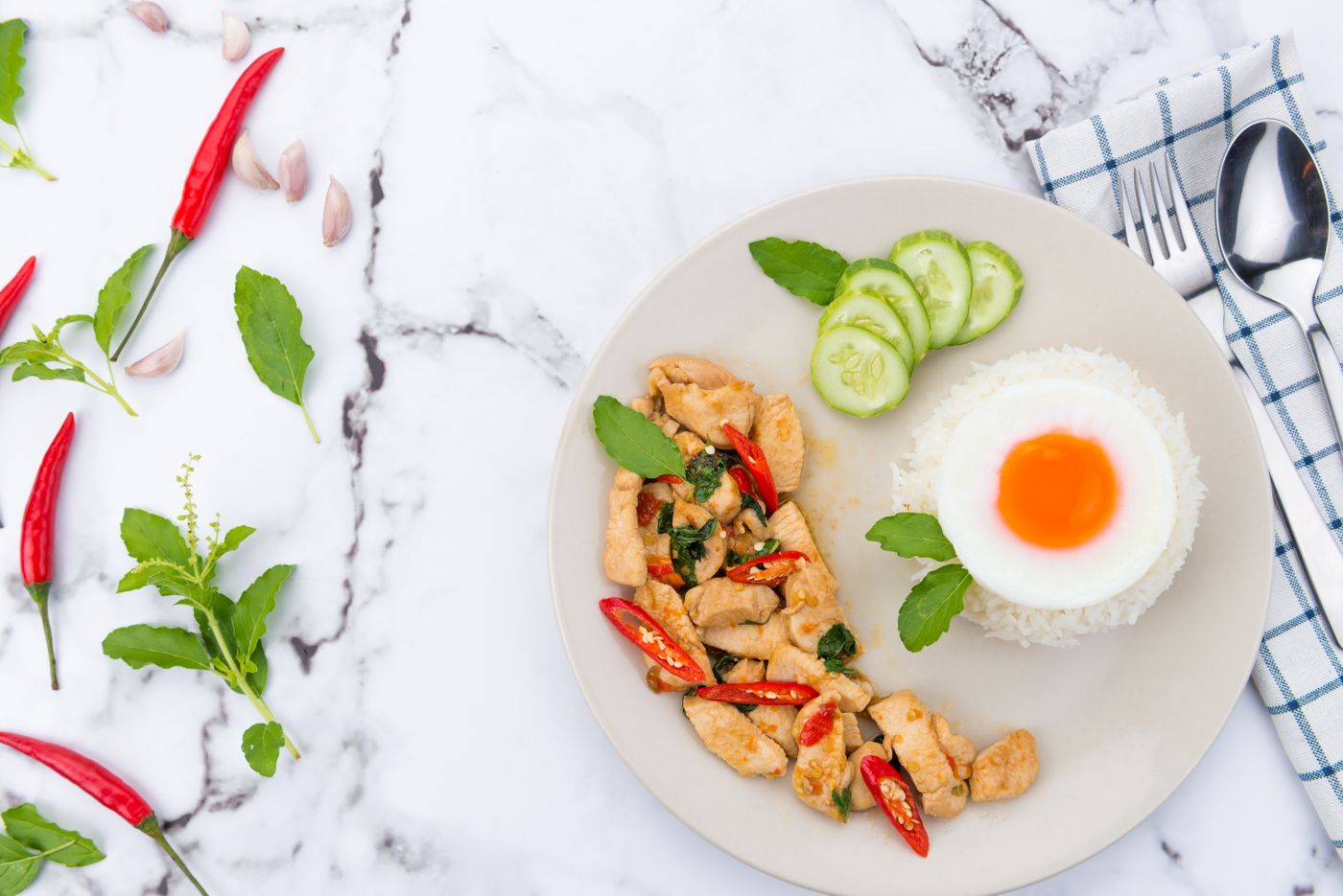 Thai basil chicken with rice and egg. chicken strips; red chili slices; basil leaves; white rice; fried egg; cucumber slices.